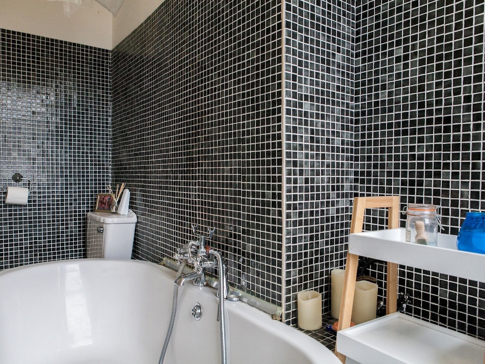 A bathroom with a white bathtub, black mosaic tiles, a small white sink, and a wooden chair.