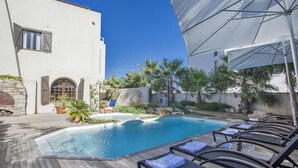 En swimmingpool med solstolar och parasoller, omgiven av en vit byggnad med välvda fönster och en stenmur.