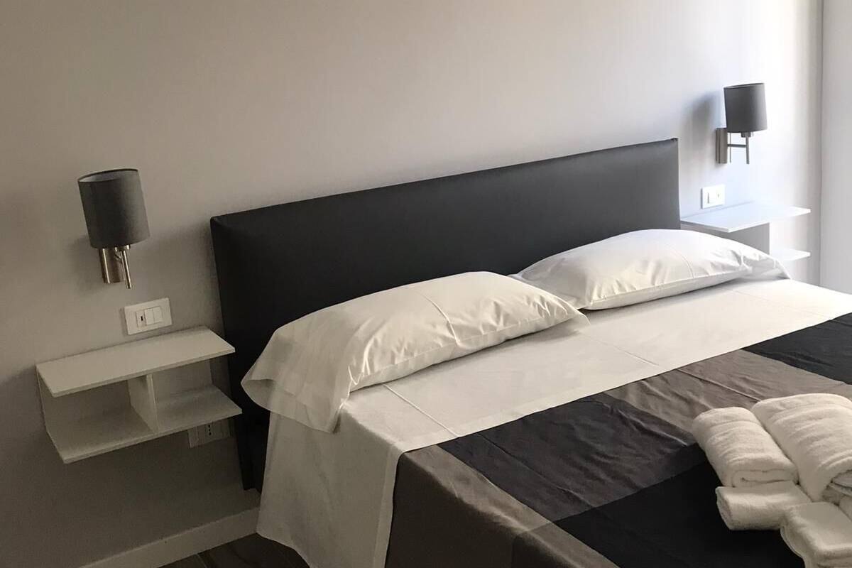 A modern hotel room with a large bed, a bedside table, a wall-mounted lamp, and a small shelf.