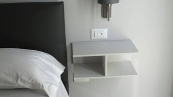 Une chambre à coucher avec un lit, une table de chevet, une lampe fixée au mur et une étagère blanche.