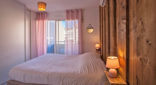 Une chambre à coucher avec un lit, une lampe de chevet, un miroir et une vue sur l’extérieur par une fenêtre avec des rideaux.