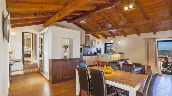 A spacious kitchen with wooden beams, a dining area with a wooden table and chairs, and a view of the outdoors through a large window.
