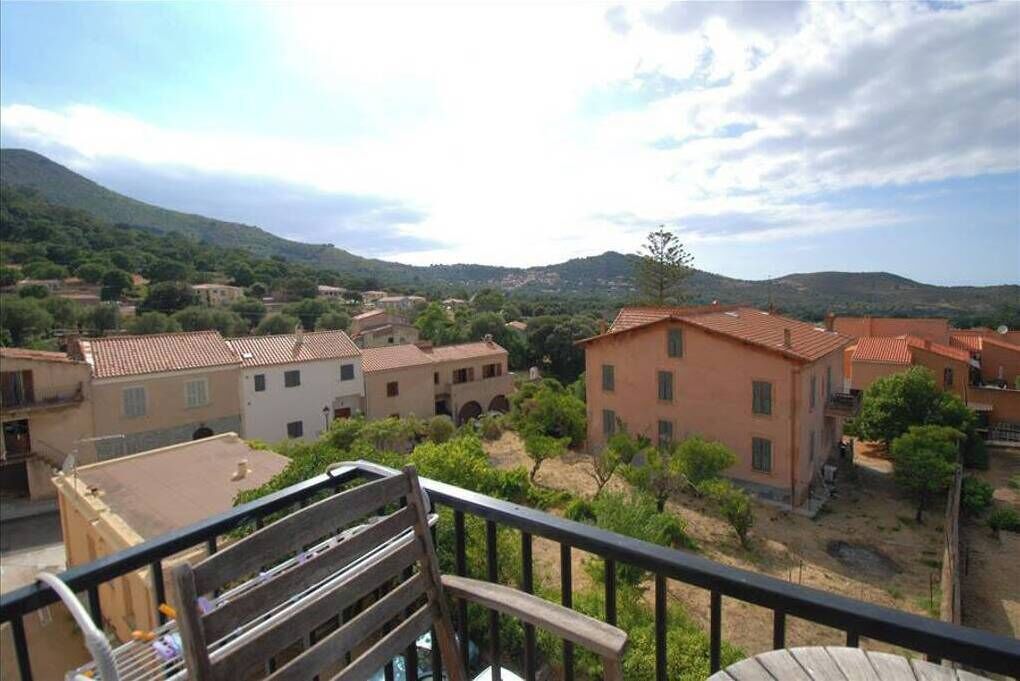 Un quartier résidentiel avec des bâtiments aux toits de tuiles, un balcon agrémenté d’une chaise en bois et un paysage montagneux en arrière-plan.