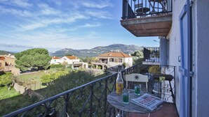 Un balcon avec un journal, une bouteille et des verres, donnant sur un village avec ses maisons et ses montagnes.