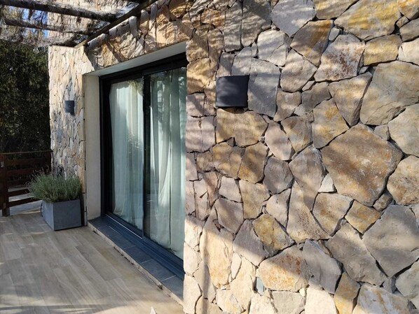 A stone wall with a glass door, a wooden deck, and a planter with shrubs.