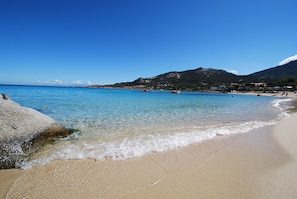 A beach with clear blue water, a rocky shoreline, and a mountain in the background.