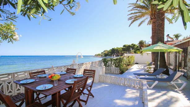 A wooden table set with plates, glasses, and a fruit bowl, overlooking a clear blue sea.