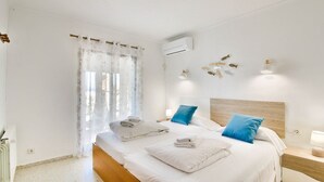 A bedroom with a bed, two pillows, a nightstand, a wall-mounted air conditioner, and a window with sheer curtains.