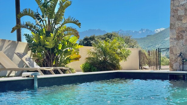 Piscina con agua cristalina, rodeada de sillas de descanso y plantas tropicales, con montañas al fondo.