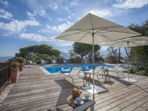 A wooden deck with a pool, lounge chairs, and an umbrella providing shade. - Villa campagne à Calvi  piscine chauffée  vue mer proche plage et centre (Calvi)