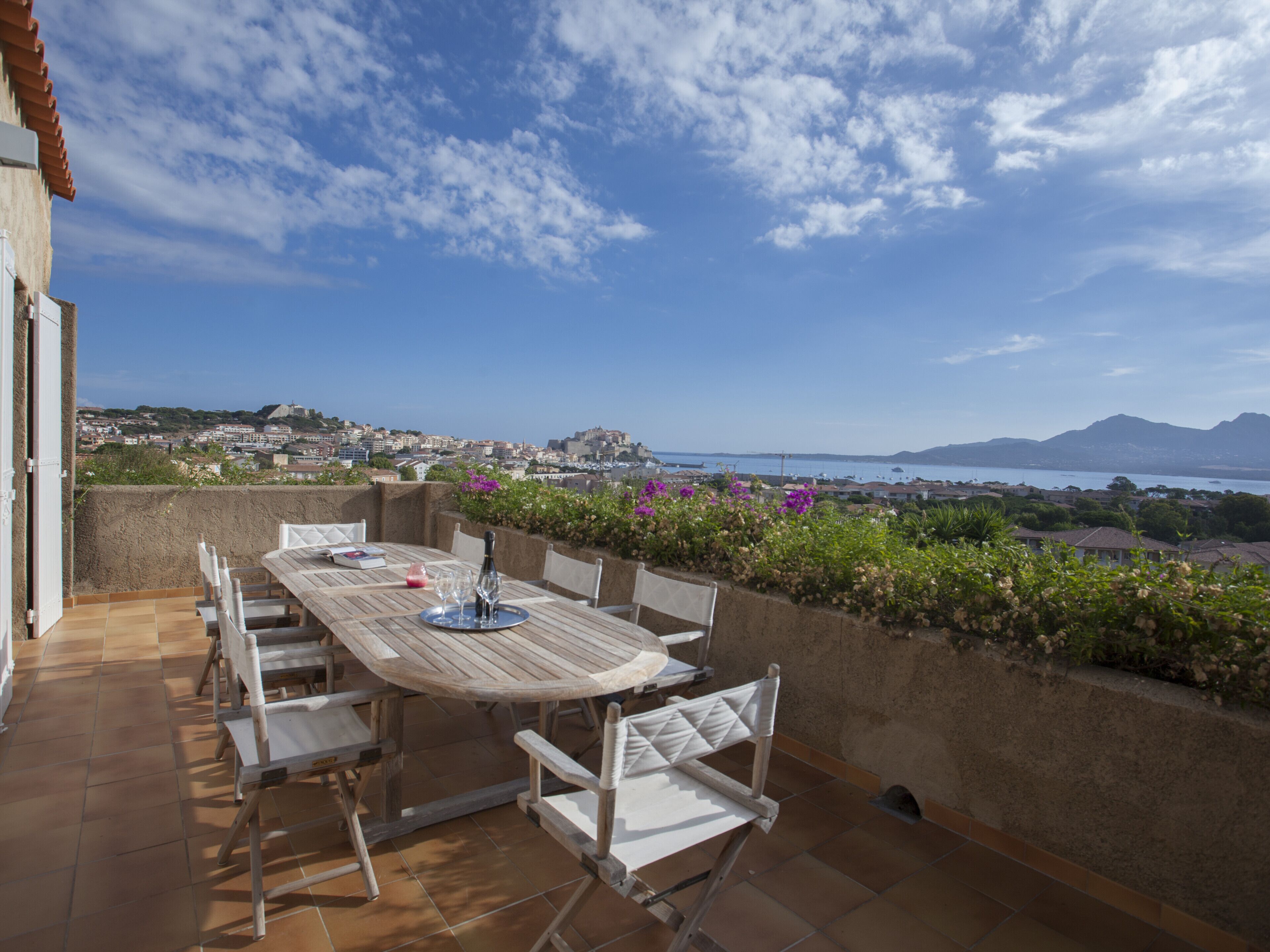 Terraza exterior con mesa y sillas de madera, con vistas a un pueblo costero y montañas.
