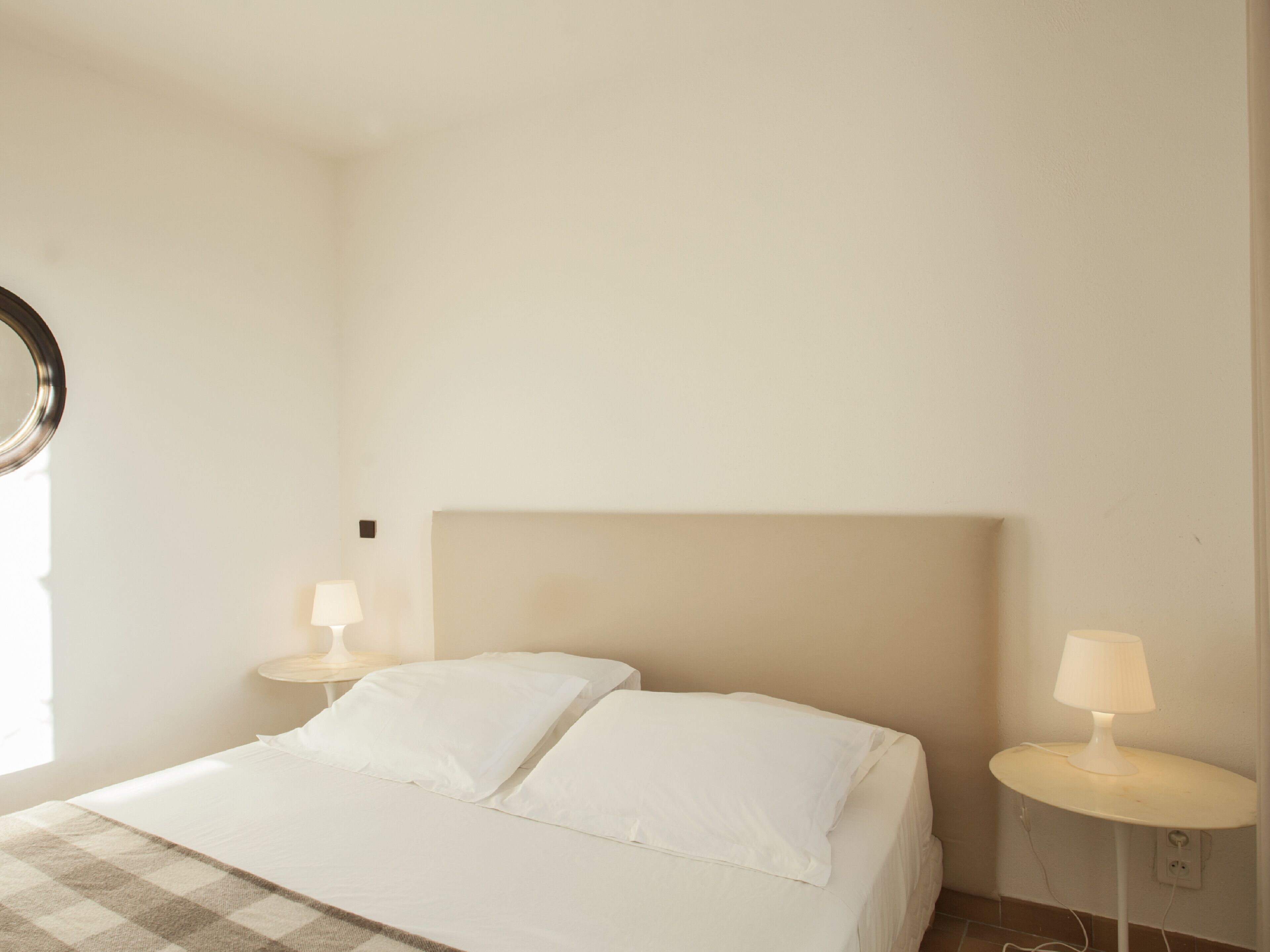 Un dormitorio minimalista con una cama grande, dos mesitas de noche con lámparas y un espejo redondo en la pared izquierda.