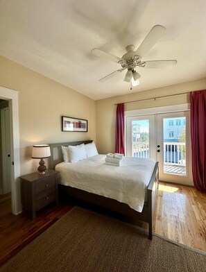 Une chambre à coucher comprenant un lit, une table de chevet, un ventilateur de plafond, une porte donnant sur un balcon et un tableau encadré au mur.