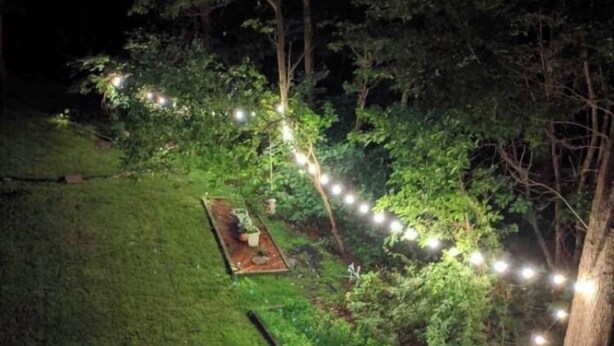 Een terras in de avond met een pad omzoomd door lichtjes, omgeven door bomen en gras.