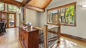 A spacious room with a high ceiling, wooden beams, a dining area with a table and chairs, a wooden staircase, and large windows offering a view of trees.