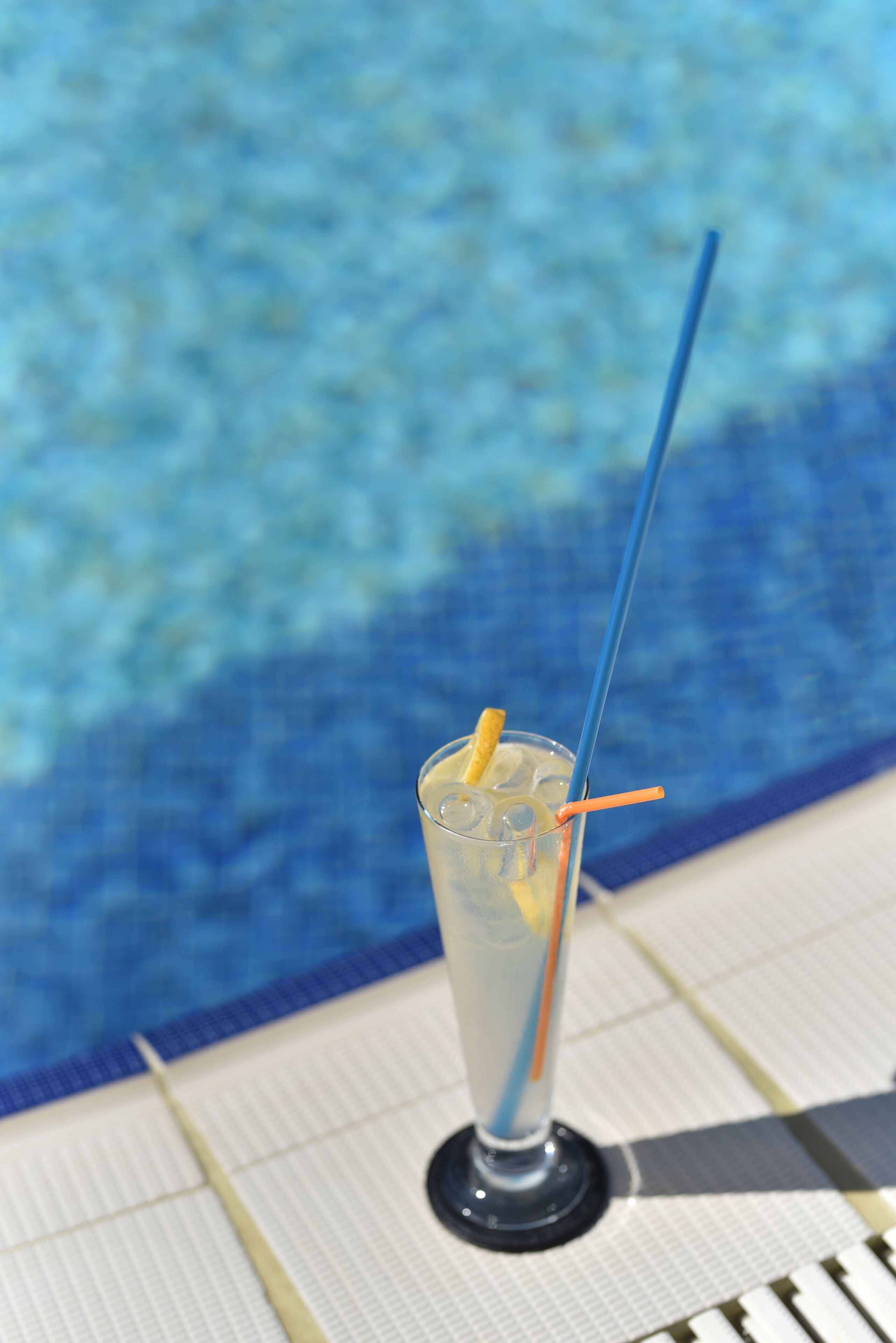 A glass of iced drink with a straw and lemon wedge on a poolside chair.
