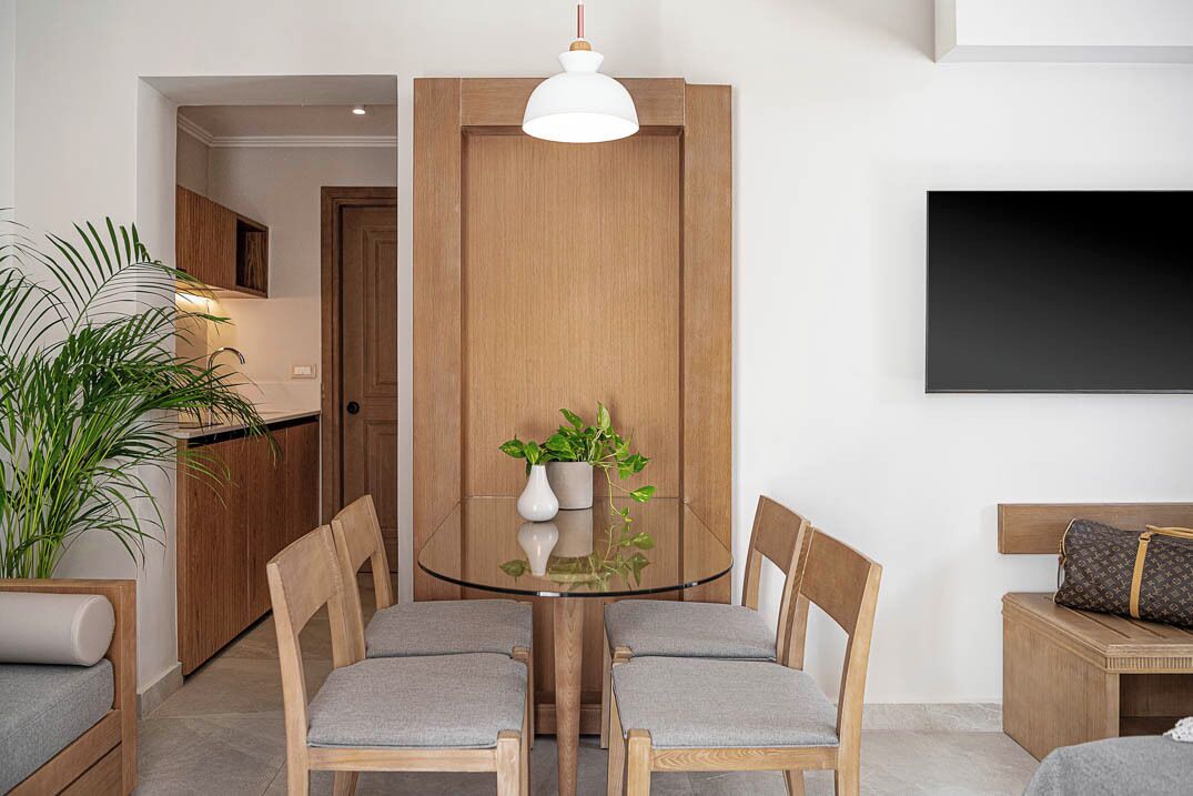 Una zona menjador moderna amb una taula de vidre, dues cadires de fusta, una planta en test, un armari de fusta i un televisor fixat a la paret.