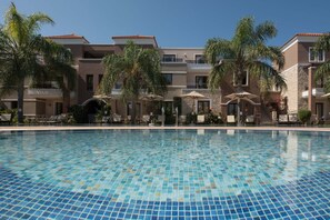 A swimming pool with tiled blue and green mosaic patterns, surrounded by multi-story buildings with balconies, palm trees, and outdoor seating areas. - Okeanis Golden Resort (Chania)