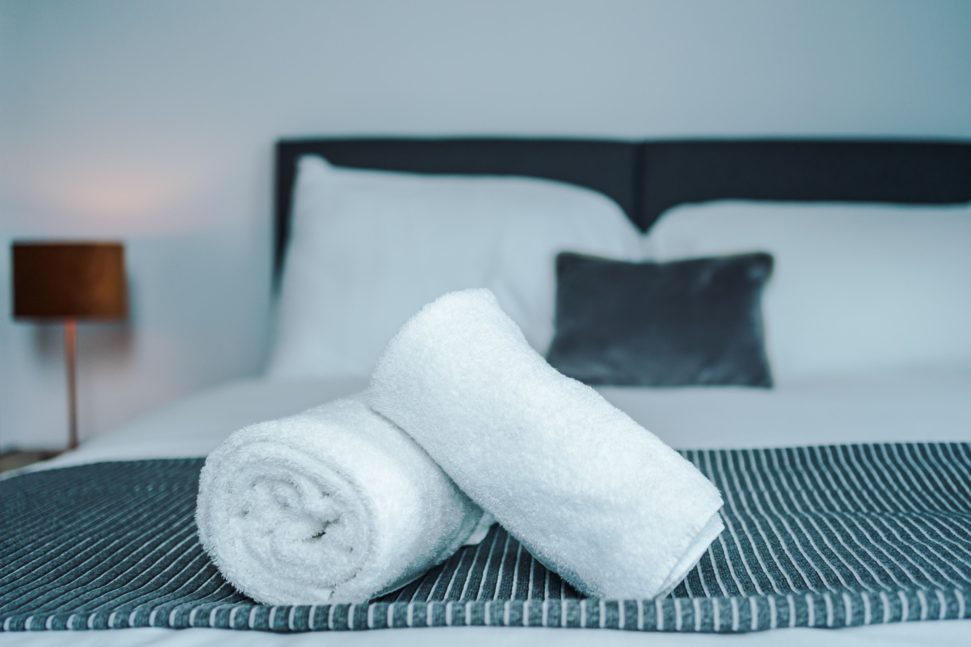 Ein ordentlich bezogenes Bett mit einer gestreiften Tagesdecke, ein aufgerolltes weißes Handtuch und ein dunkles Kopfstück.