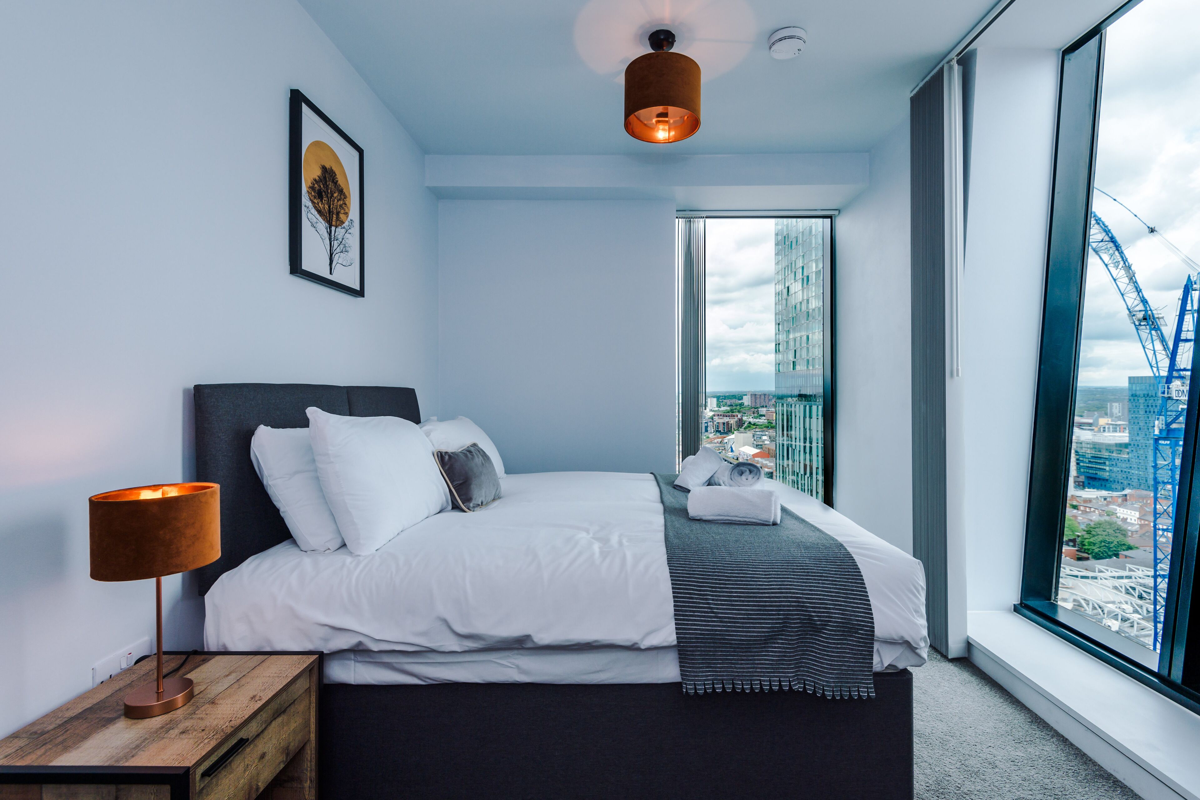 Une chambre d’hôtel moderne avec un grand lit, une table de chevet avec une lampe, un tableau encadré au mur et une vue sur le paysage urbain par une grande fenêtre.