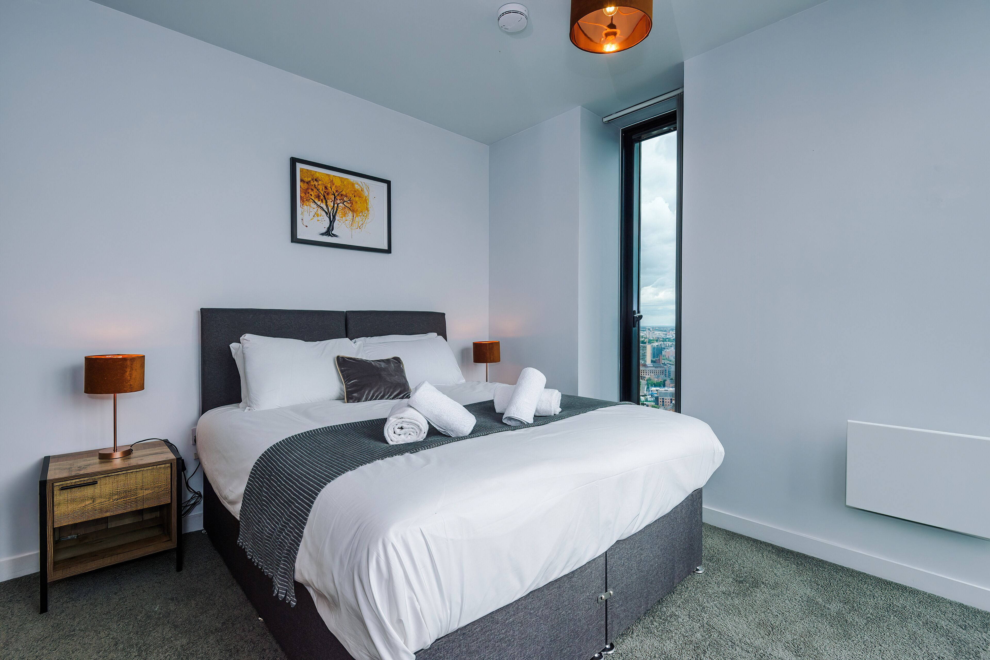 Une chambre d’hôtel moderne avec un grand lit, deux tables de chevet avec des lampes, une vue sur la ville à travers une grande fenêtre et une œuvre d’art encadrée au mur.
