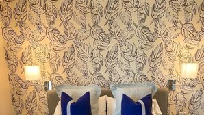 A neatly made bed with a white duvet and blue pillows, a tufted ottoman, and a wall with a leafy pattern. - The Lofts at Paso Market Walk - The Elderberry Room (Paso Robles)