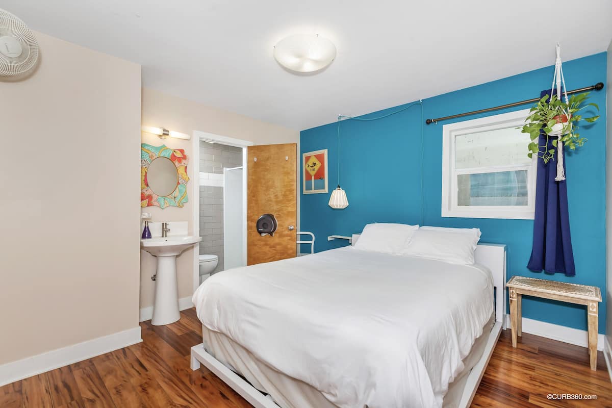 Een slaapkamer met een bed, een nachtkastje, een hangende plant, een raam met gordijnen, een wandlamp en een deur naar een badkamer.