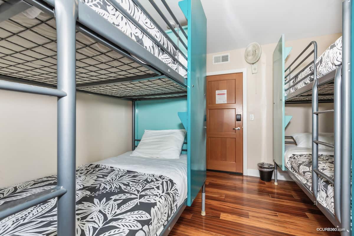 Een slaapkamer met stapelbed, een houten deur, een prullenbak en een ventilator aan de muur.