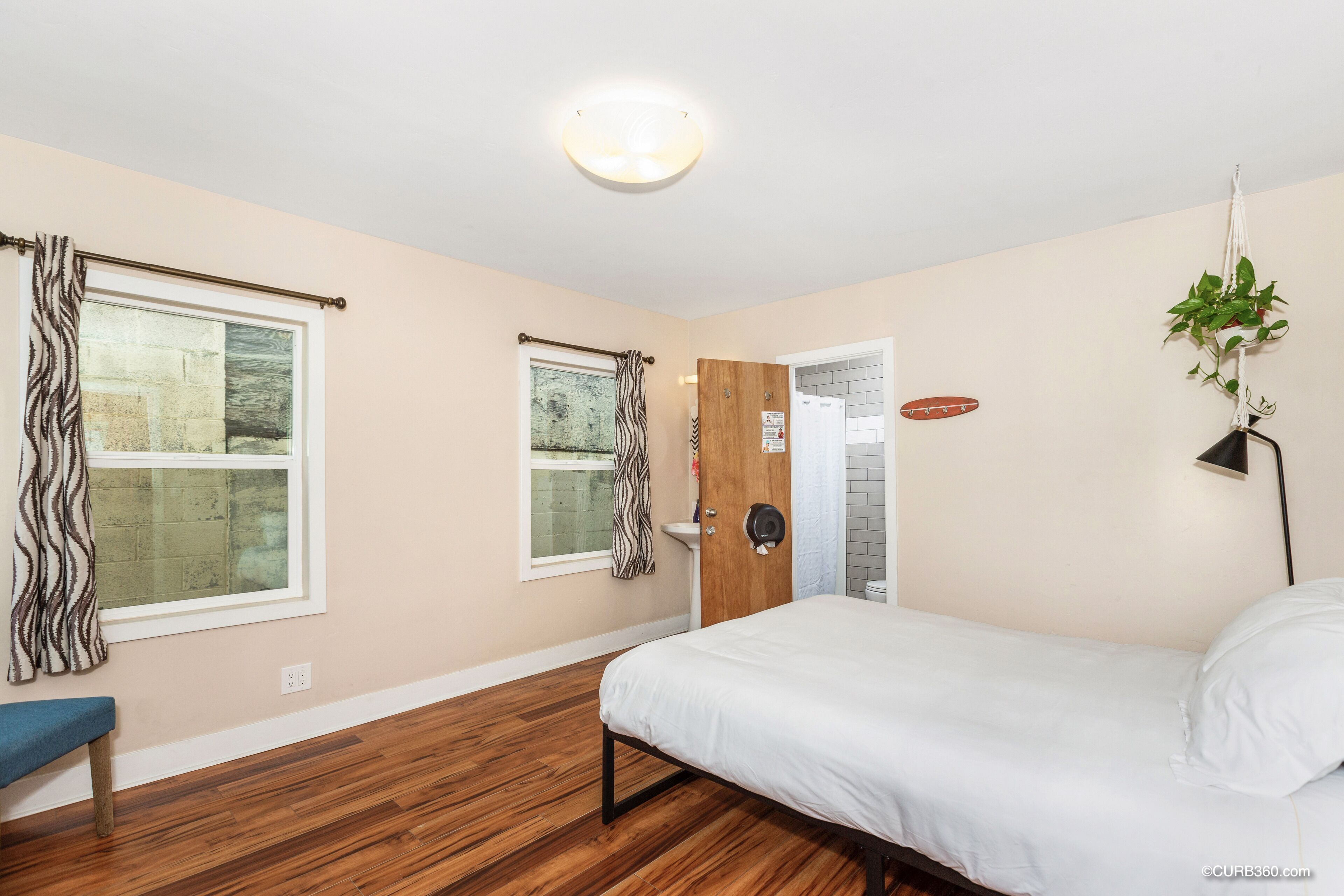 Un dormitorio con cama, suelo de madera, un banco, ventana con cortinas, una puerta y una planta colgante.