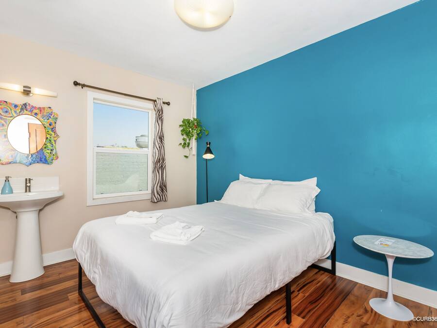 A bedroom with a bed, a round mirror, a white sink, a window with a zebra curtain, a potted plant, and a small round table.