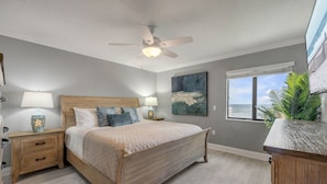 Un dormitorio con cama de madera, mesitas de noche, ventilador de techo, ventana con vistas y un cuadro en la pared.