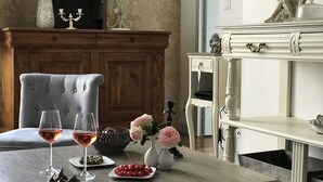 A dining area with a wooden cabinet, a white sideboard, and a grey upholstered chair. There are two wine glasses on a table, a vase with flowers, and a basket with fruits.