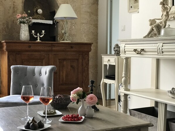 Un comedor con un armario de madera, un aparador blanco y una silla tapizada en gris. Hay dos copas de vino sobre una mesa, un florero con flores y una cesta con frutas.