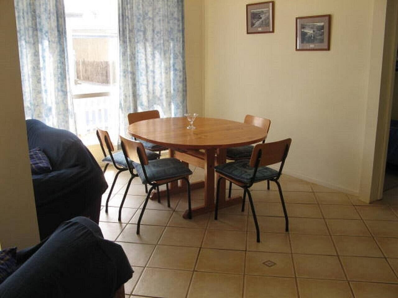Un comedor con una mesa redonda de madera y cuatro sillas, una ventana con cortinas, y dos cuadros en la pared.