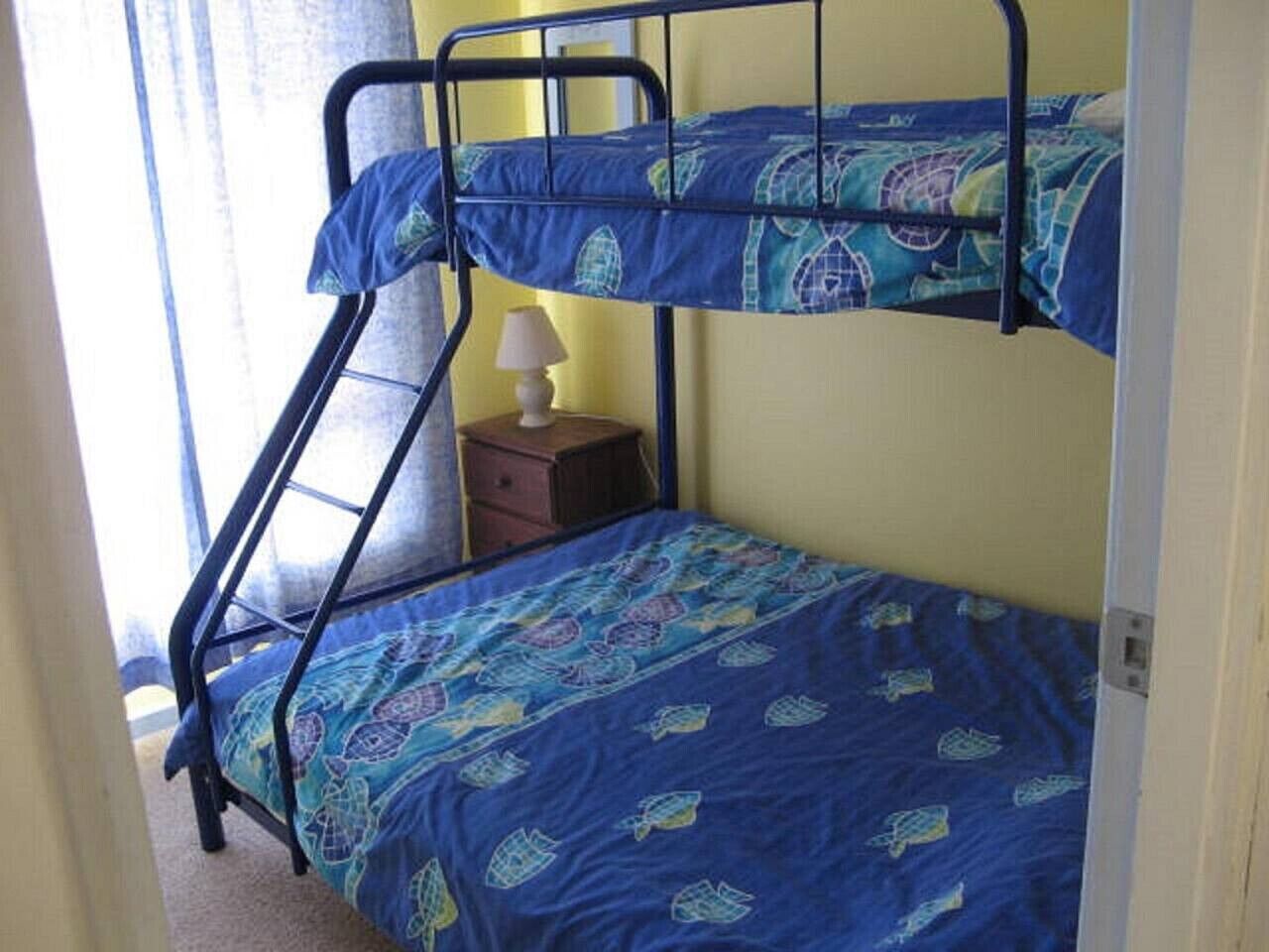 A bunk bed with a patterned blue bedspread, a small wooden nightstand with a lamp, and a window with curtains.