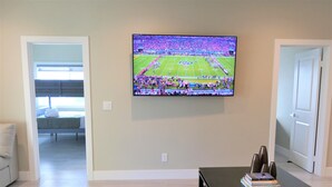 Uma sala de estar com televisão exibindo uma partida de futebol, uma mesa de centro preta com livros e controles remotos, e um sofá.