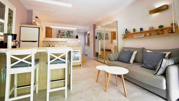 A modern kitchen with a central island, a dining area with white chairs, and a grey sofa with cushions.