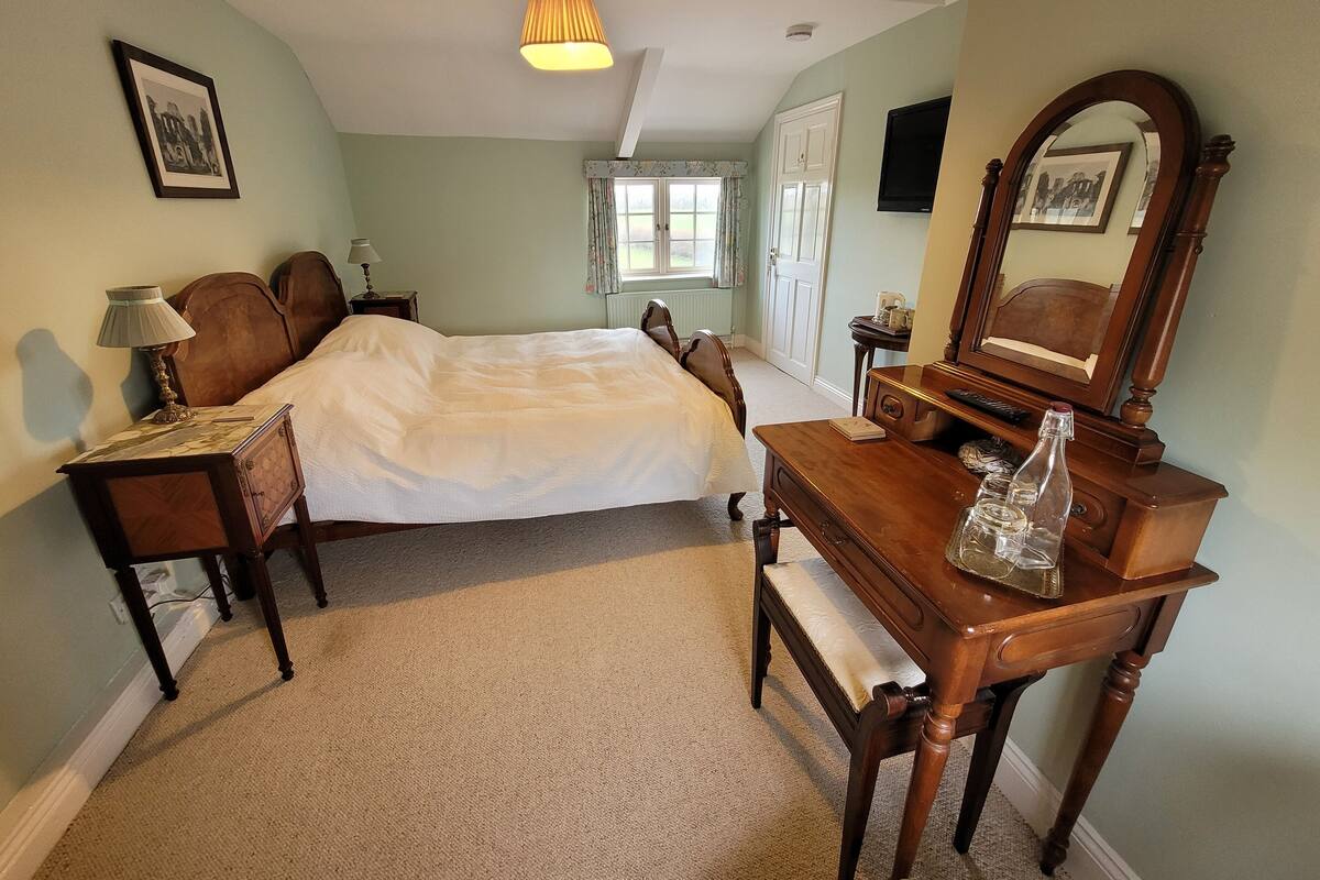 Un dormitorio con una cama, un aparador de madera, una mesita de noche, una lámpara, un espejo y una ventana con cortinas.
