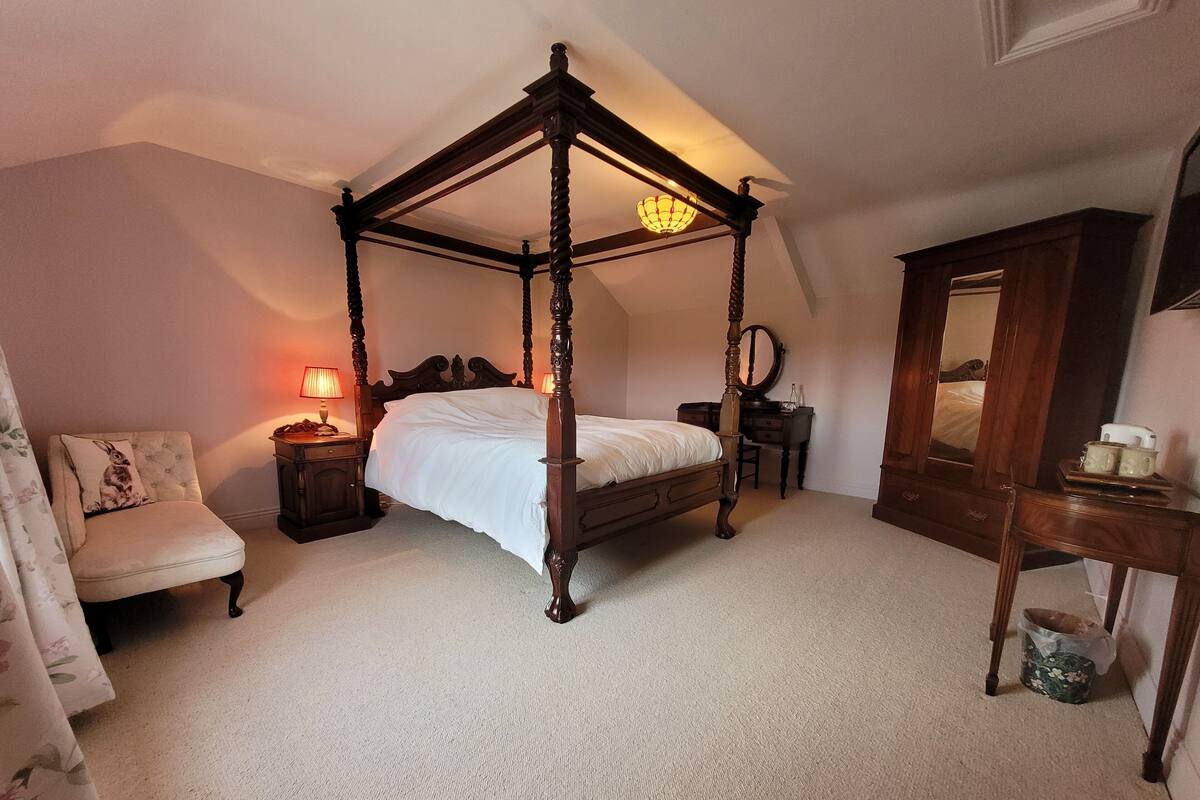 Una cama con dosel en un dormitorio con mesita de noche, silla, cómoda y espejo.