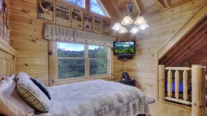 A cozy wooden cabin bedroom with a bed, pillows, a television, and a large window offering a scenic view. - AMAZING GATED COMMUNITY - YOUR OWN PRIVATE SAUNA & HOT TUB ON BACK PORCH (Sevierville)