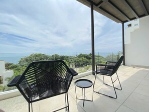 Relax on the balcony with two comfortable chairs and a small table, taking in the serene views of the lush landscape and distant sea.