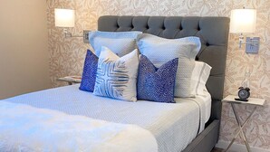 Una cama bien hecha con cabecera gris, ropa de cama blanca y almohadillas decorativas. Hay dos mesitas de noche con lámparas y un reloj pequeño. La habitación tiene papel tapiz estampado y una alfombra con diseño en el suelo.