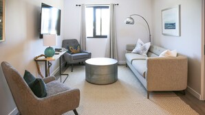Una sala de estar moderna con un sofá gris, una mesa de centro redonda, un sillón gris, una mesita de noche con una lámpara y un televisor montado en la pared.
