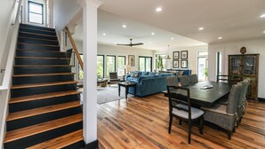 Ein geräumiges Wohnzimmer mit einer dunklen Holztreppe, einer blauen Sofagarnitur, einem Esstisch mit Stühlen und einem Deckenventilator.