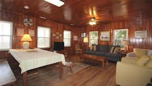 Ein gemütliches Zimmer mit Holzwänden und -decke, einem Esstisch mit weißem Tischlein, einer Couch und einem Fernseher.