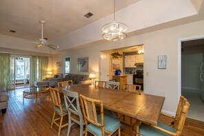 Un vaste espace de salle à manger avec une grande table en bois et des chaises en osier, un ventilateur de plafond et une cuisine visible à travers une porte ouverte.