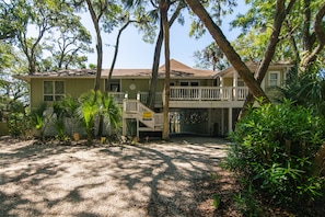 A two-story house with a balcony, surrounded by trees and a gravel driveway.