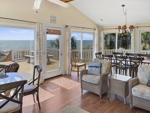Une pièce spacieuse dotée d’une grande porte-fenêtre coulissante qui donne sur une terrasse offrant une vue sur l’océan. À l’intérieur, on trouve des fauteuils en osier et une table, et un lustre est suspendu au plafond.