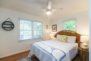 Une chambre à coucher avec un lit, deux fenêtres, un ventilateur de plafond et une étagère fixée au mur.