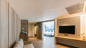 A modern hotel room with a sofa, a TV mounted on the wall, and a view of the city through large windows.